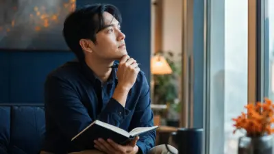 Seorang mahasiswa duduk dekat jendela menulis jurnal untuk refleksi diri dan menjaga kesehatan mental di lingkungan kampus